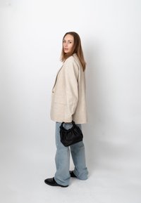 Frau trägt einen beigen Blazer und hellblaue Jeans, hält eine schwarze, geraffte Handtasche und steht vor einem schlichten weißen Hintergrund.