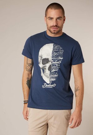 Homme aux cheveux courts et tatouages, portant un t-shirt bleu marine avec un grand motif demi-crâne et du texte blanc, un pantalon beige et des bracelets.