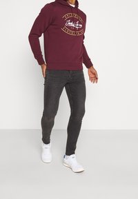 Bordeauxroter Kapuzenpullover mit weißem und gelbem Logo, kombiniert mit schwarzen Jeans und weißen Turnschuhen. Verfügt über eine klassische Passform und weichen Stoff.