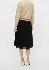 Jupe plissée noire avec un design fluide, fabriquée en tissu léger. Associée à un haut beige ajusté à manches longues et des chaussures à talons blocs beiges.