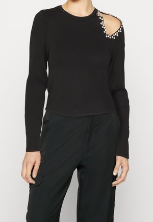 Haut noir à manches longues avec une découpe embellie sur l'épaule droite, associé à un pantalon noir.