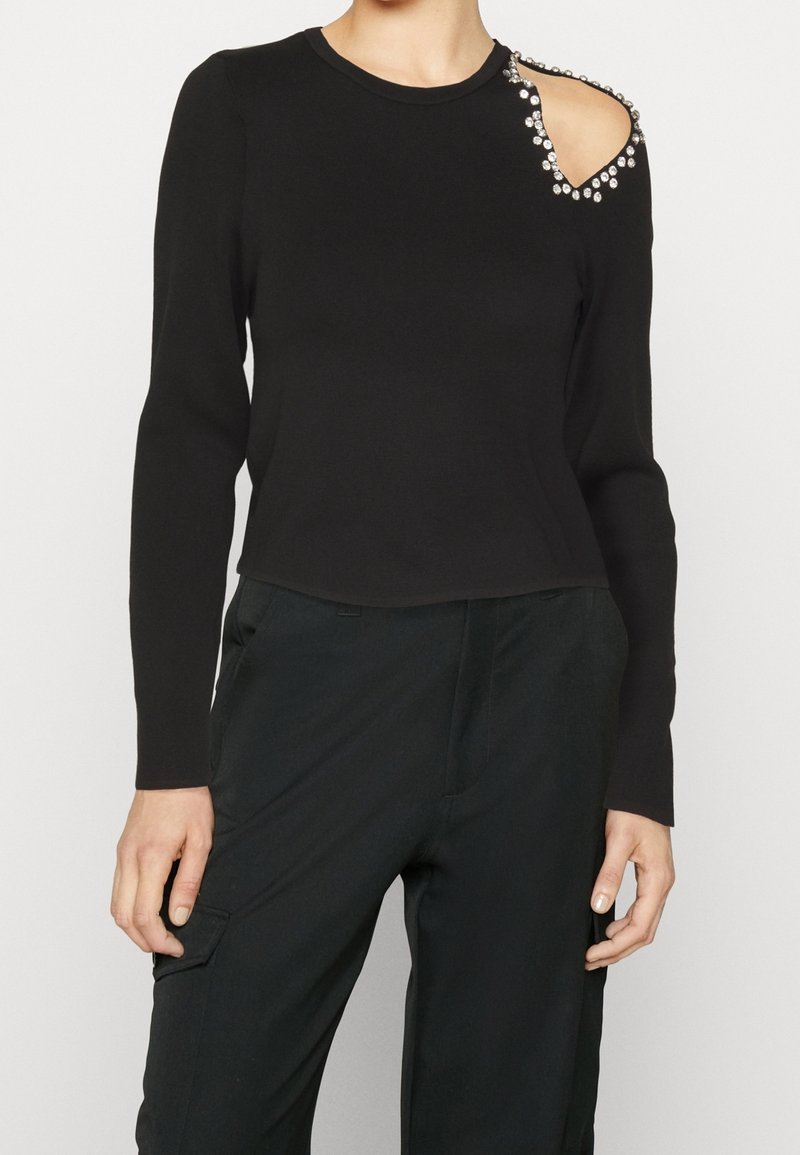 Haut noir à manches longues avec une découpe embellie sur l'épaule droite, associé à un pantalon noir.
