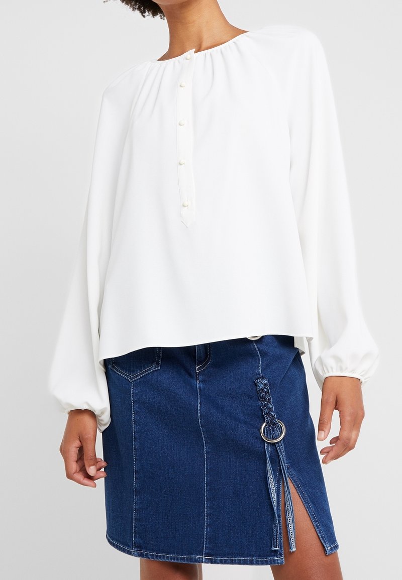 Blusa bianca con maniche lunghe e a sbuffo, dal collo rotondo, caratterizzata da cinque piccoli bottoni frontali. Abbinata a una gonna di jeans.