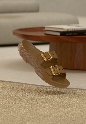 Bruine instapsandaal met twee verstelbare gouden gespen zwevend boven een geweven beige tapijt, nabij een ronde houten salontafel met gestapelde boeken.