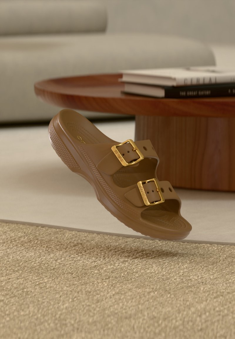 Sandale marron enfilable avec deux boucles dorées réglables flottant au-dessus d’un tapis beige tissé, près d’une table basse ronde en bois avec des livres empilés.