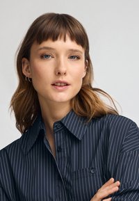 Granatowa koszula w paski z guzikami i kołnierzykiem, z kieszonką i błyszczącymi guzikami, noszona przez modela z włosami do ramion.