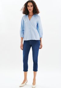 Blusa di cotone azzurro chiaro con scollatura a V e maniche a palloncino di lunghezza metà, con chiusura a bottoni e dettagli ricamati. Abbinata a jeans capri blu scuro.