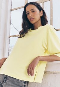 Gelbes kurzärmeliges T-Shirt aus weicher Baumwolle mit Rundhalsausschnitt, seitlichen Schlitzen und lockerer Passform. Kombiniert mit grauen Denim-Shorts.