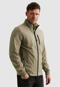 Muž s krátkymi hnedými vlasmi, oblečený v svetlo béžovej bunde na zips s vysokým golierom a čiernych džínsach, stojaci s jednou rukou v vrecku.