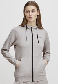 Svijetlosmeđi zip-up dukserica izrađena od mekanog materijala. Ima kapuljaču, crni patentni zatvarač, logo na prsima i dva bočna džepa.