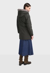 Ciemnozielona wodoodporna kurtka z kapturem wyłożonym futrem, w połączeniu z niebieską spódnicą dżinsową i brązowymi wysokimi botkami, widok z tyłu.