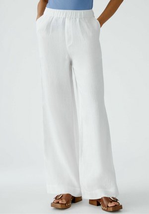 Femme portant un pantalon blanc ample avec poches et des sandales à plateforme marron, les mains dans les poches.