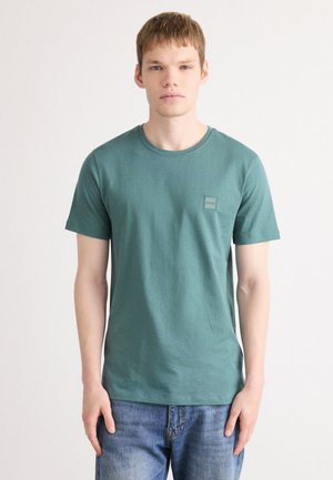 Grønn bomull t-skjorte med rund hals og korte ermer, med en liten firkantet logo på venstre brystområde. Kombinert med blå jeans.