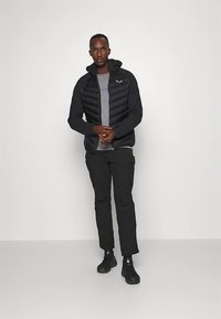 Svart vadderad jacka med huva, grå tröja under och svarta byxor; slät textur och sportig passform, kompletterad med svarta skor.