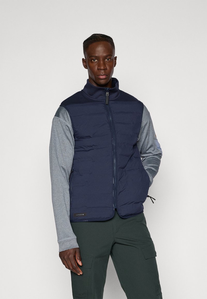 Gilet imbottito blu con zip frontale, alto collo e maniche lunghe grigie. Presenta una texture liscia e orlo elasticizzato con toppa logo.