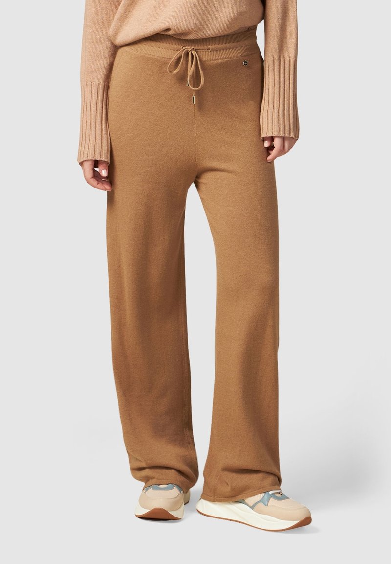 Personne portant un pantalon large en maille couleur chameau avec taille à cordon, un pull beige et des baskets beige et blanches sur fond clair.