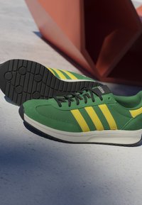 Green and yellow Adidas sneaker with black laces and black sole resting on a light-colored surface near a red geometric object.