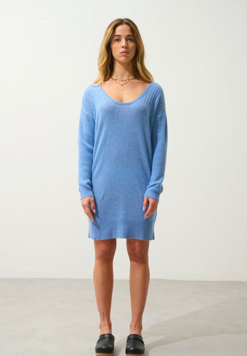 Světle modré pletené šaty v uvolněném střihu, s ribbovaným límcem a rukávy a hladkou strukturou, doplněné černými dřeváky na šedé podlaze.