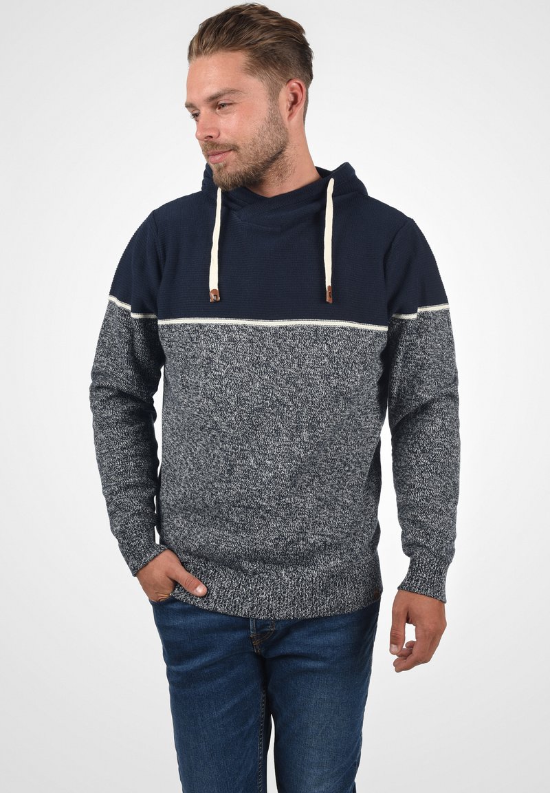 Sweat à capuche avec une partie supérieure bleu marine et une partie inférieure grise texturée. Il présente des cordons de serrage crème et un ourlet côtelé pour s'ajuster.
