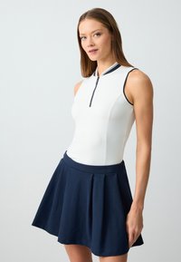 Witte mouwloze top met een ritskraag en een marineblauwe geplooide rok. De outfit heeft een gladde textuur en contrasterende accenten.