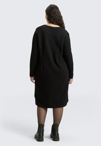 Zwart sweater jurkje van een textuurstof, met lange mouwen en een brede halslijn, tot halverwege de kuit. Gecombineerd met enkelhoge laarzen en transparante panty's.