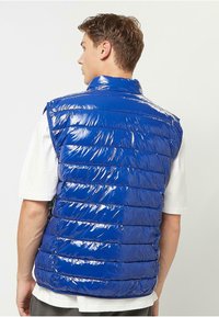 Blå polstret vest, skinnende tekstur, ærmeløs design, kviltet mønster med vandrette linjer, rund krave, båret over en hvid skjorte.