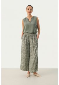 Mouwloze olijfgroene blouse met een uitgesneden halslijn, gecombineerd met losse, gestreepte wijde broek in tinten groen en crème.