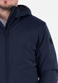 Marineblaue Kapuzenjacke mit glatter, matter Oberfläche. Verfügt über einen Reißverschluss vorne und ein rundes schwarzes Logo an der linken Ärmels.