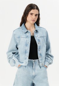 Veste en denim bleu clair au design classique, avec un devant à boutons, des poches poitrine et une coupe décontractée. Portée sur un haut noir, associée à un jean clair.