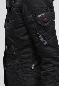 Veste matelassée noire avec fermeture éclair devant, détails à boutons, multiples poches et tissu texturé. Présente un logo sur la manche.