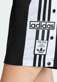 Čierno-biele športové oblečenie s tromi vertikálnymi pruhmi, vyšívaným logom a gombíkovým detailom na boku. Textúrovaná látka.