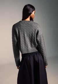 Sivi pulover kratke dolžine z vzorcem pletenja v krog, z okroglim izrezom, kombiniran s tekočo temno vijolično krilo. Mehka tekstura, sproščen kroj in rebrast rob.