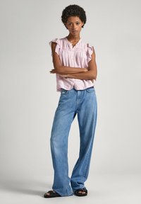 Chemise boutonnière à manches courtes, rose clair, avec des manches à volants ; portée avec un jean en denim bleu ample et des sandales noires.