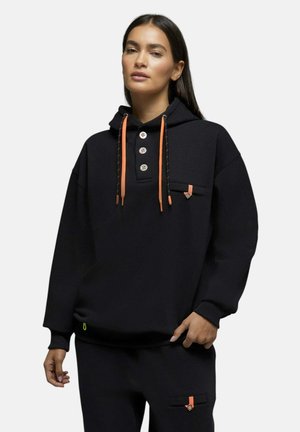 Femme portant un sweat à capuche noir avec des cordons orange et un pantalon noir assorti avec des poches accentuées orange, posant devant un fond uni.