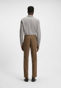 Camicia button-up grigia chiara a maniche lunghe, abbinata a pantaloni beige su misura. Tessuto liscio, colletto classico e dettaglio con piega sul retro.