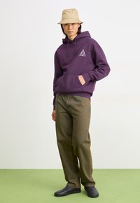 Sudadera morada con bolsillo frontal y logo en forma de triángulo, combinada con pantalones verdes oliva texturizados y zapatos negros sin cordones; sombrero tipo cubo beige.