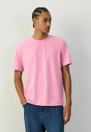 Jeune homme avec des tresses cornrows portant un t-shirt rose uni à manches courtes et un jean bleu, debout avec une main dans la poche.
