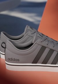 Zapatillas Adidas grises con rayas negras y logo, suelas blancas, mostradas de lado y ligeramente desde arriba sobre un fondo azul y naranja.