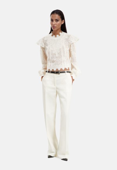 Blouse en dentelle blanche avec des épaules volantées, assortie à un pantalon blanc taille haute. Une ceinture noire accentue la tenue. Tissu léger et doux.