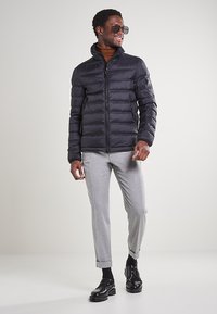 Giubbotto a sacco nero con cuciture orizzontali, colletto alto e chiusura con zip. Indossato sopra un dolcevita marrone e pantaloni tapered grigio chiaro.