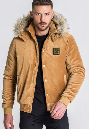 Chaqueta de ante color marrón claro con capucha de piel sintética, botones a presión y un logo bordado en negro y dorado en el lado izquierdo del pecho. Textura suave, diseño casual.