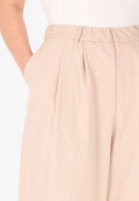 Pantaloni beige a vita alta realizzati in un tessuto liscio e leggero, con pieghe sul davanti e tasche laterali. Dettagli delle cuciture lungo la vita.