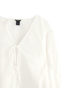 Blusa bianca con scollo a V profondo, dettaglio a fiocco al collo e maniche lunghe arricciate. Il tessuto testurizzato aggiunge una sottile profondità al design.
