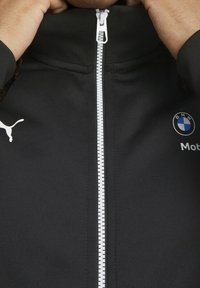 Černá zipová bunda vyrobená z elastické tkaniny, s bílým zipem, logem Puma a odznakem BMW na prsou. Hladká textura.