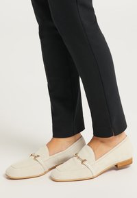 Mocasini din suede bej cu un toc mic din lemn, având accente din metal auriu și vârful rotunjit, asortați cu pantaloni negri croiți.