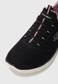 Juodi Skechers sportbačiai su rožiniais akcentais, megztas viršus, elastingos bungee raišteliai ir balta pado dalis, kampu fotografuota artima išvaizda.