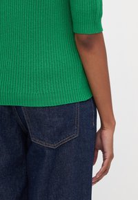 Grön ribbad stickad tröja med korta ärmar, kombinerad med mörkblå denimjeans med detalj på bakfickan.