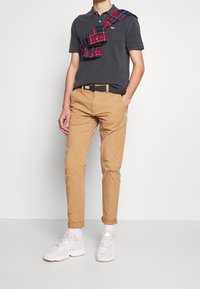 Grijze polo, beige chinos en witte sneakers. Geruit shirt over de schouder geknoopt. Eenvoudig ontwerp, slanke pasvorm, effen kleuren en opgerolde broekspijpen.