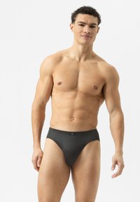 Costume da bagno nero a slip con un motivo geometrico, realizzato in tessuto elasticizzato. Design a vita alta e una morbida fascia in vita. Modello in posa in piedi.