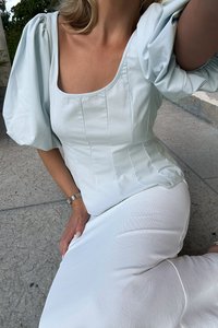 Helles blaues Top mit Puffärmeln, einem quadratischen Ausschnitt und vertikalen Nähten, kombiniert mit einem taillierten weißen Rock. Der Stoff wirkt weich und glatt.